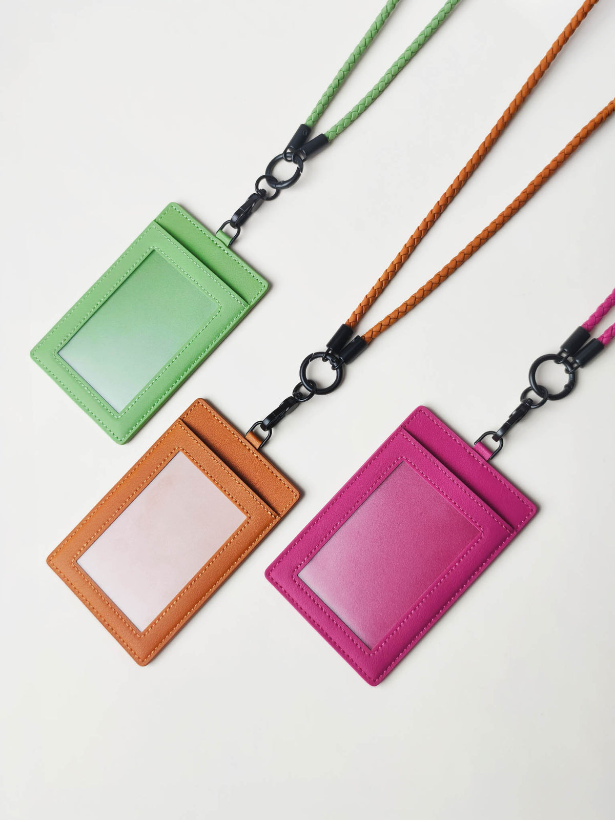 Three sicora vegan leather ID badge holders with braided loop  lanyards in green, orange, and pink, arranged diagonally on a light off-white background.