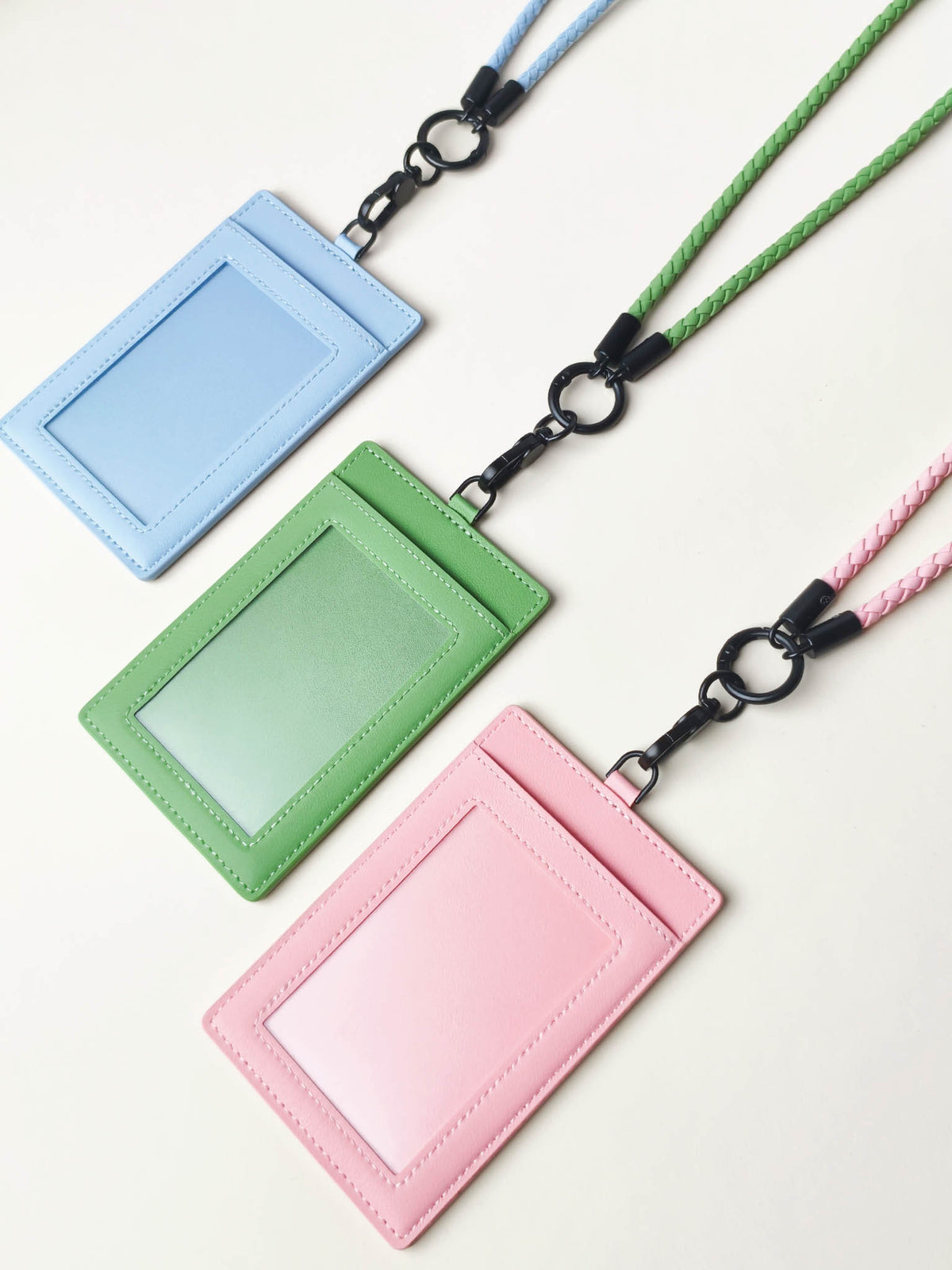 Three sicora vegan leather ID badge holders with braided lanyards in light blue, green, and pink, arranged diagonally on a light cream background.
