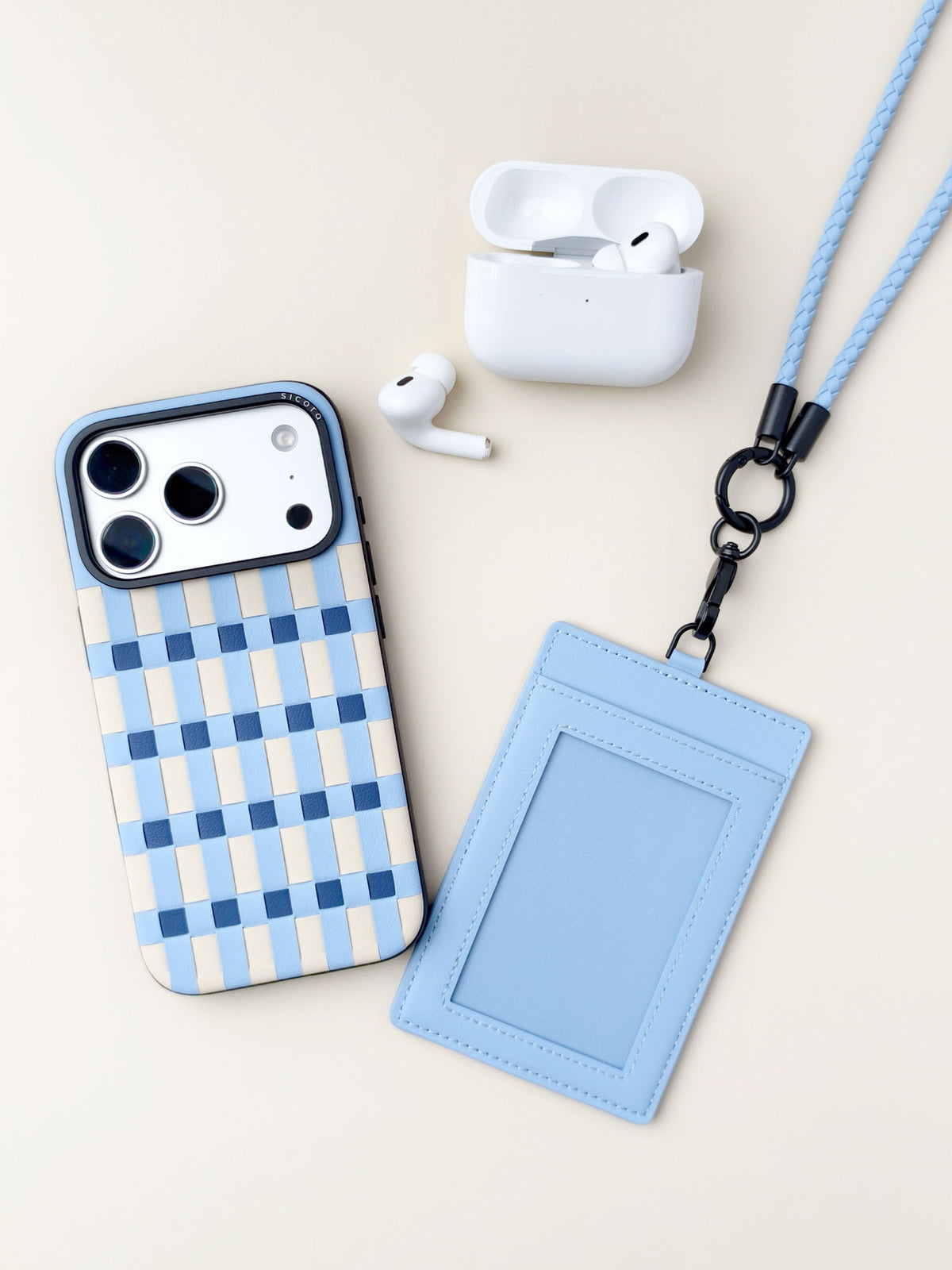 A flat-lay product shot featuring sicora vegan leather iPhone 18 / 17 pro case, pixel case, a matching light blue vegan leather ID badge holder with a black carabiner and braided lanyard, a white AirPods charging case,.