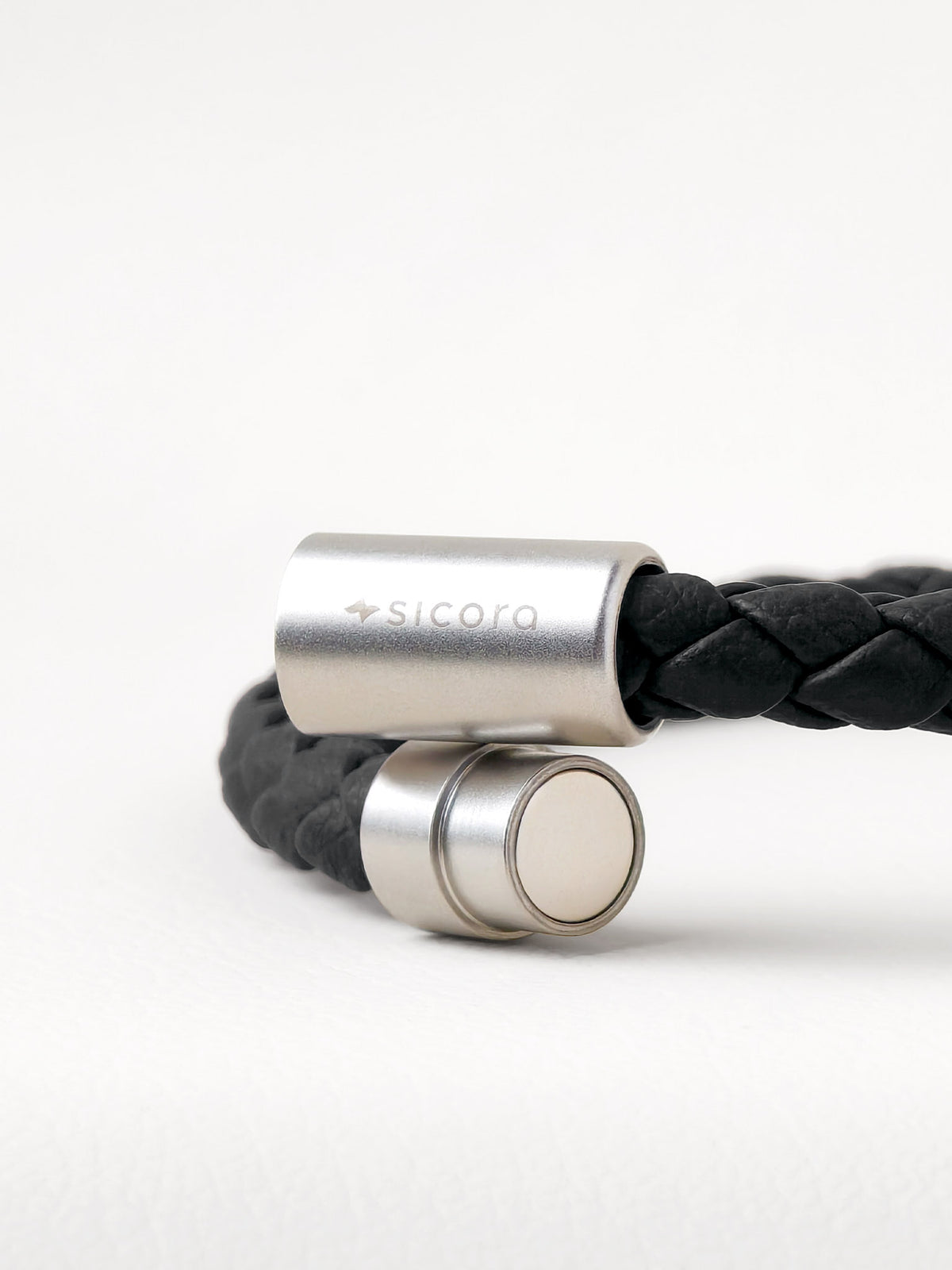 Close-up of a sicora black braided leather bracelet with stainless steel magnetic end caps, one of which is engraved with the brand name 'sicora'.