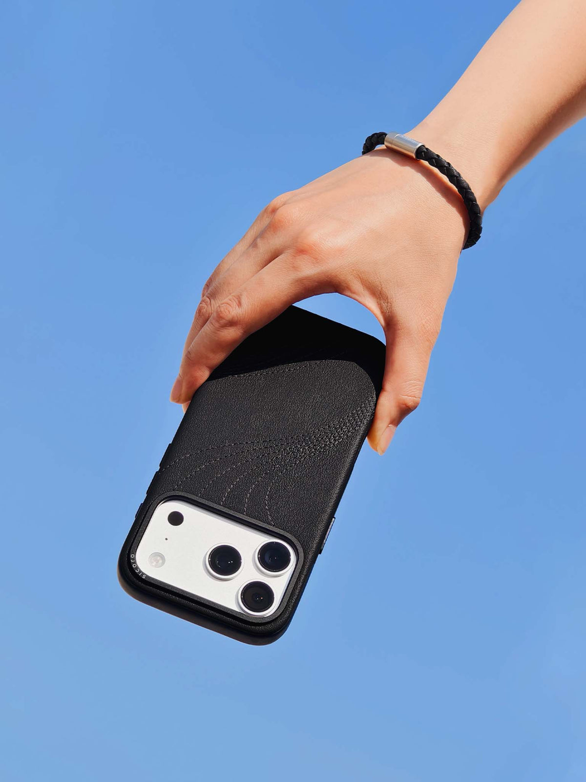 Hand holding a black sicora vegan leather iPhone case with a sicora braided leather bracelet on the wrist, clear blue sky background.