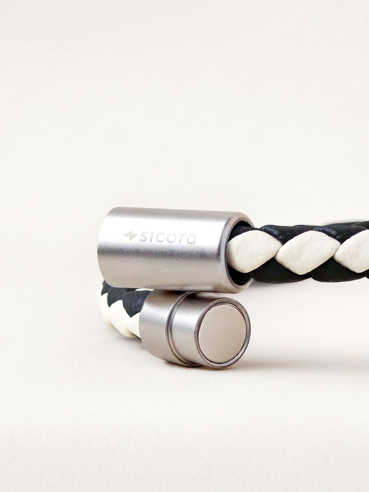 Close-up of a sicora cream black two-tone braided leather bracelet with stainless steel magnetic end caps, one of which is engraved with the brand name 'sicora'.