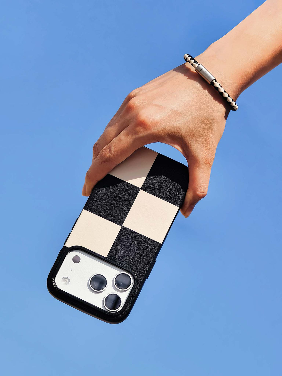 Hand holding a black cream sicora vegan leather iPhone case with a matching sicora braided leather bracelet on the wrist, clear blue sky background.