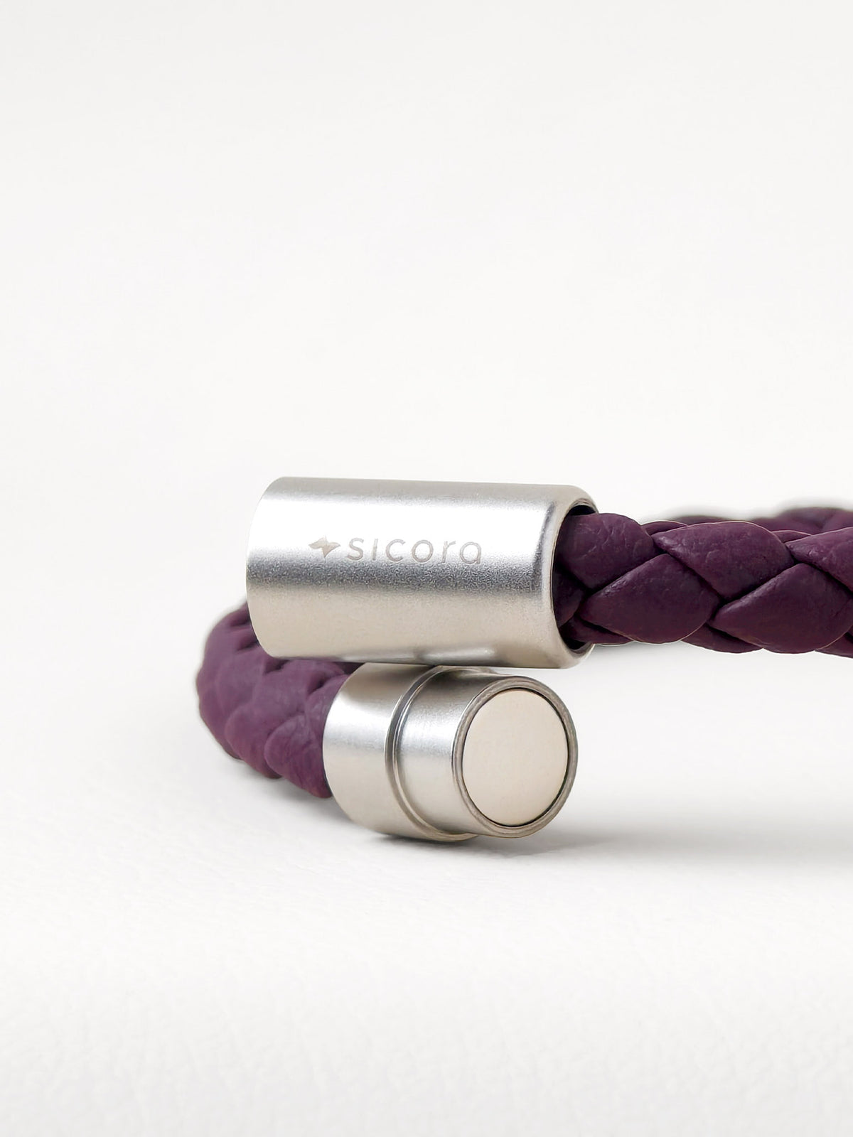 Close-up of a sicora dark cherry purple braided leather bracelet with stainless steel magnetic end caps, one of which is engraved with the brand name 'sicora'.