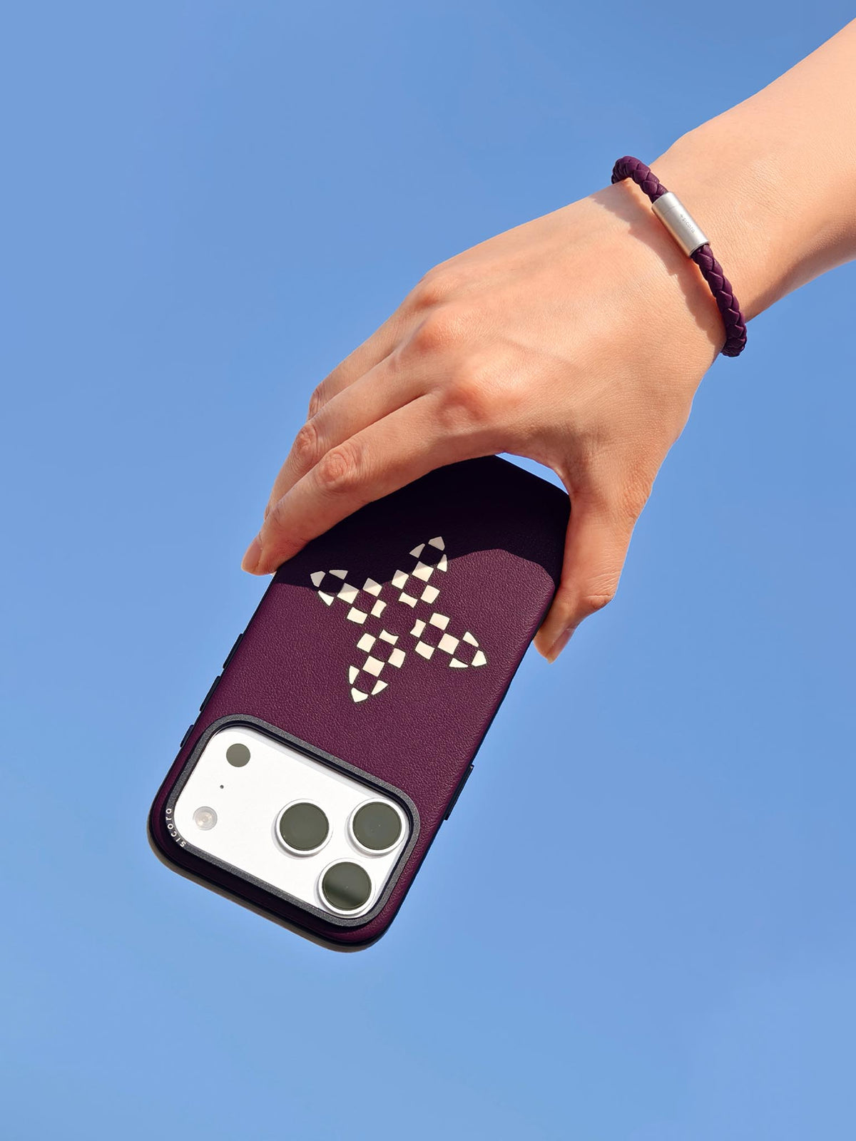 Hand holding a dark cherry purple sicora vegan leather iPhone case with a sicora braided leather bracelet on the wrist, clear blue sky background.