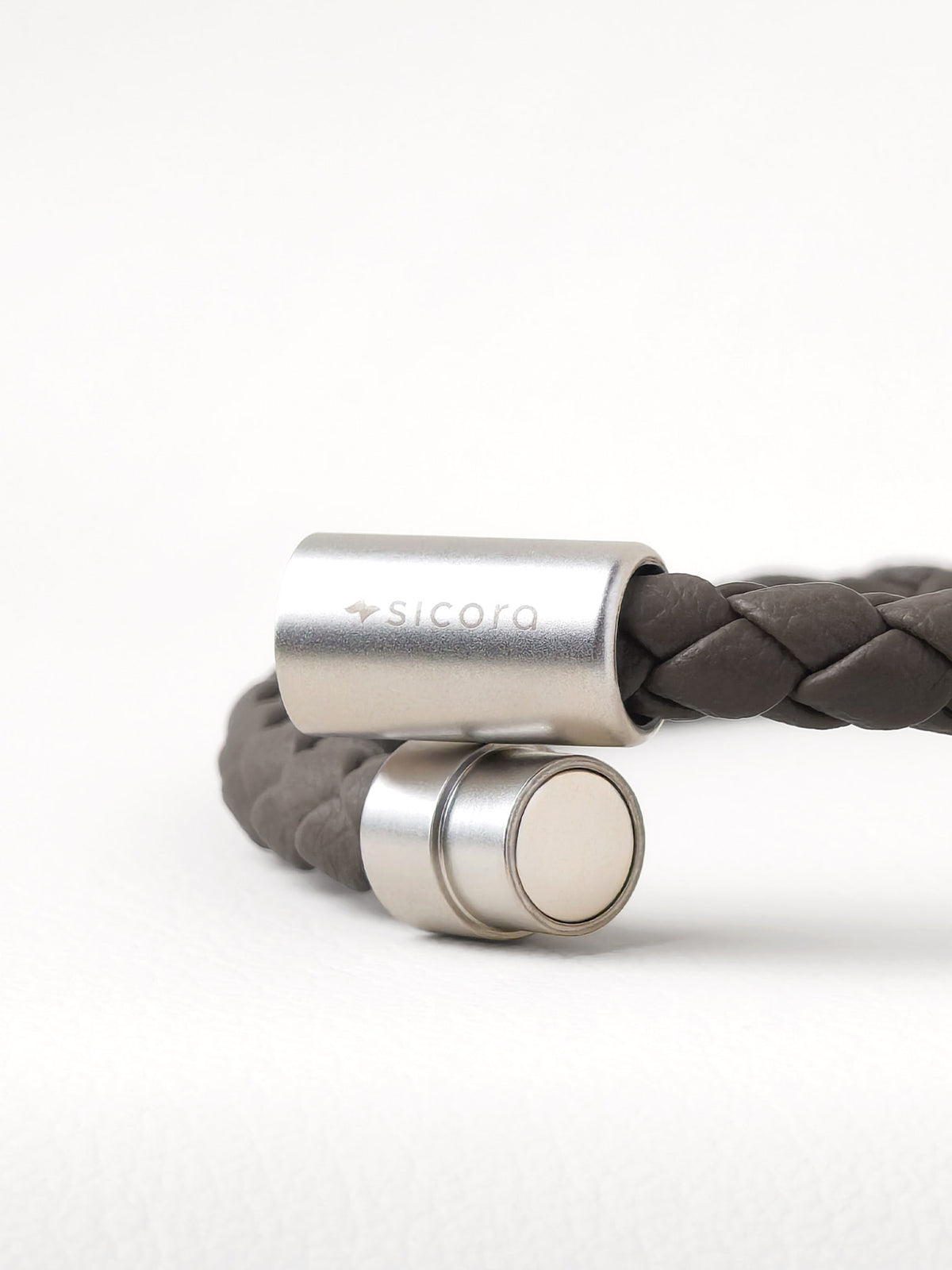 Close-up of a sicora gray / grey braided leather bracelet with stainless steel magnetic end caps, one of which is engraved with the brand name 'sicora'.