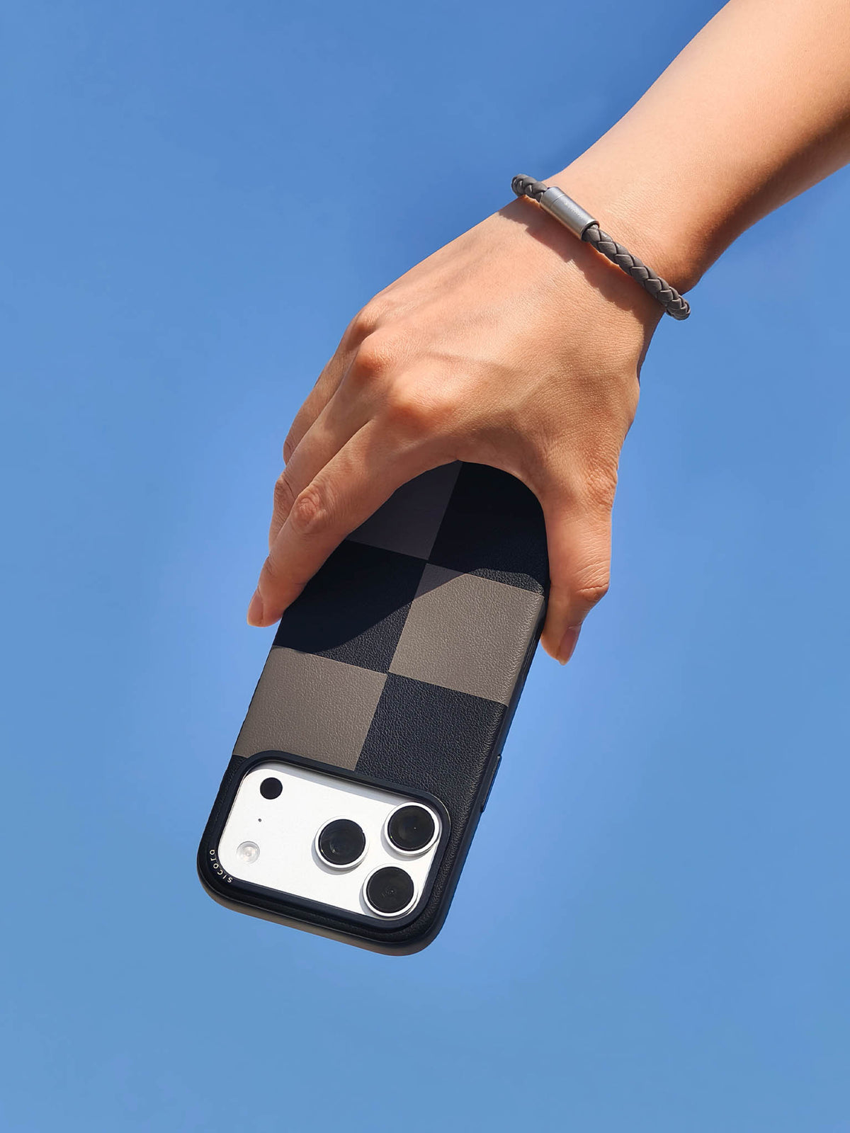 Hand holding a black gray sicora vegan leather iPhone case with a gray sicora braided leather bracelet on the wrist, clear blue sky background.