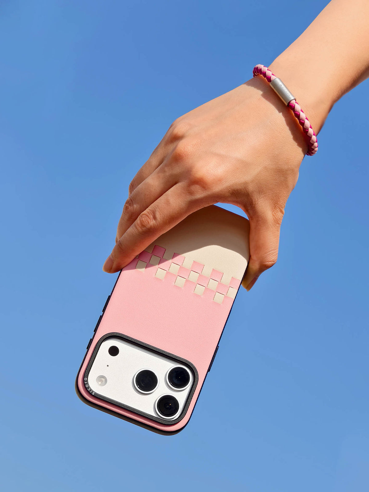 Hand holding a pink cream sicora vegan leather iPhone case with a pink magenta sicora braided leather bracelet on the wrist, clear blue sky background.
