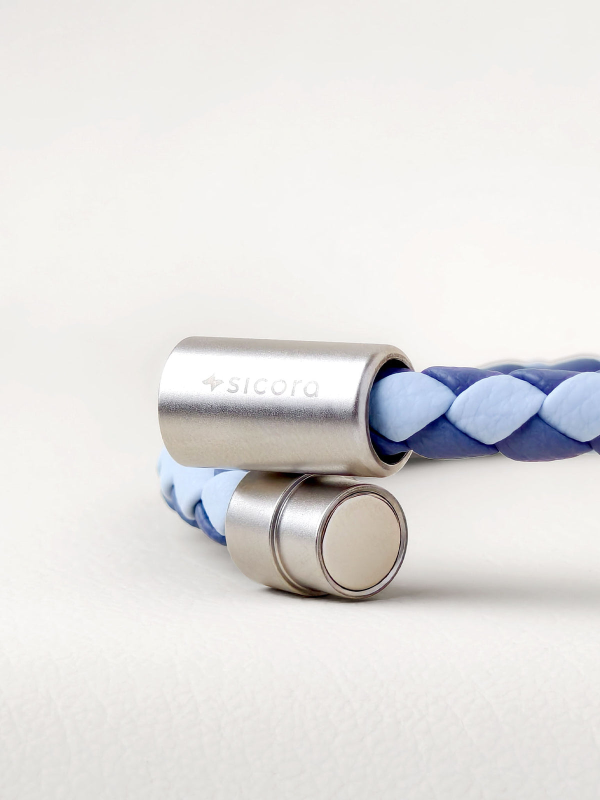 Close-up of a sicora sky blue and shadow blue two-tone braided leather bracelet with stainless steel magnetic end caps, one of which is engraved with the brand name 'sicora'.