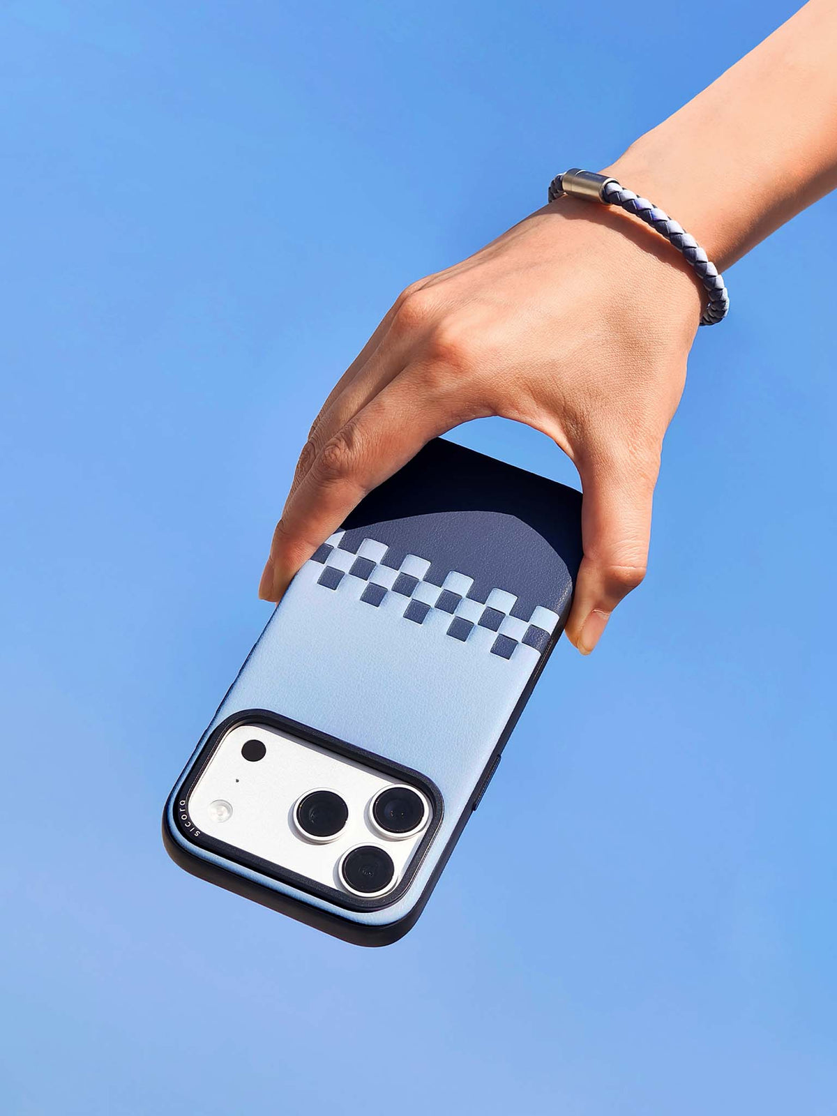 Hand holding a sky shadow blue sicora vegan leather iPhone case with a matching sicora braided leather bracelet on the wrist, clear blue sky background.