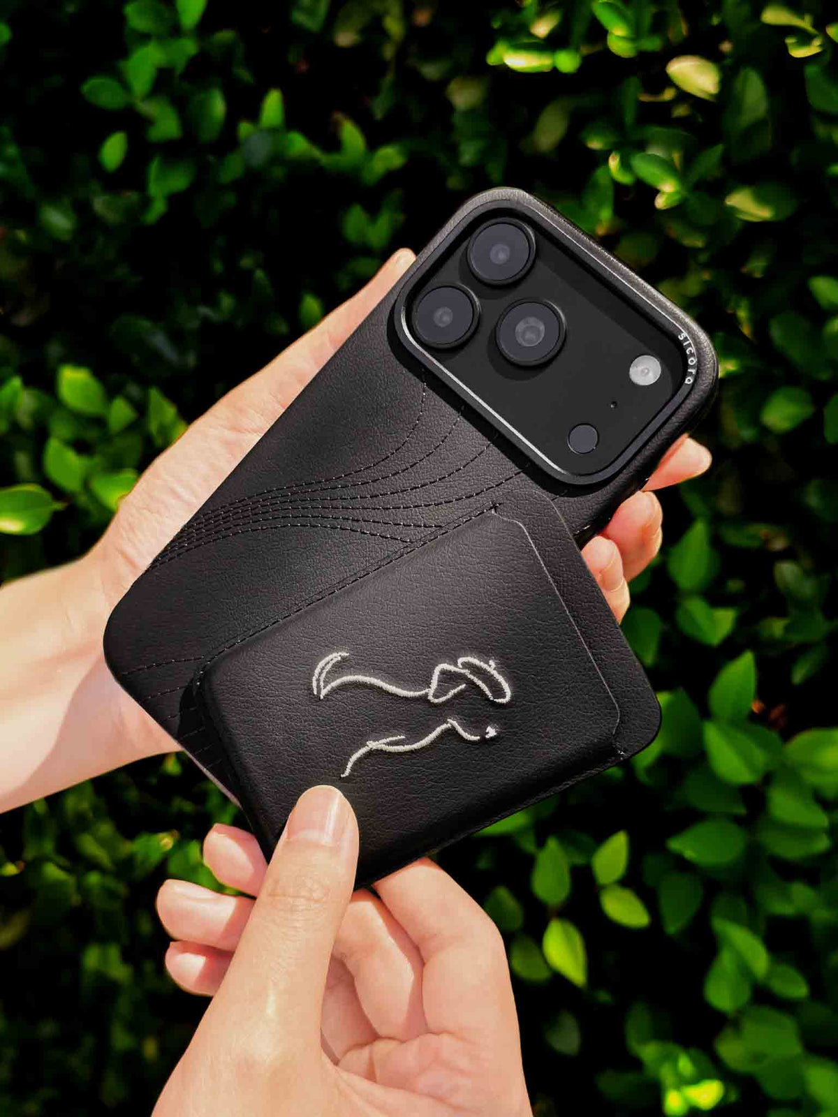 Hands holding a sicora vegan leather graphic magnetic card wallet in black with cream white embroidered dog line-drawing & a sicora iPhone 18 pro case