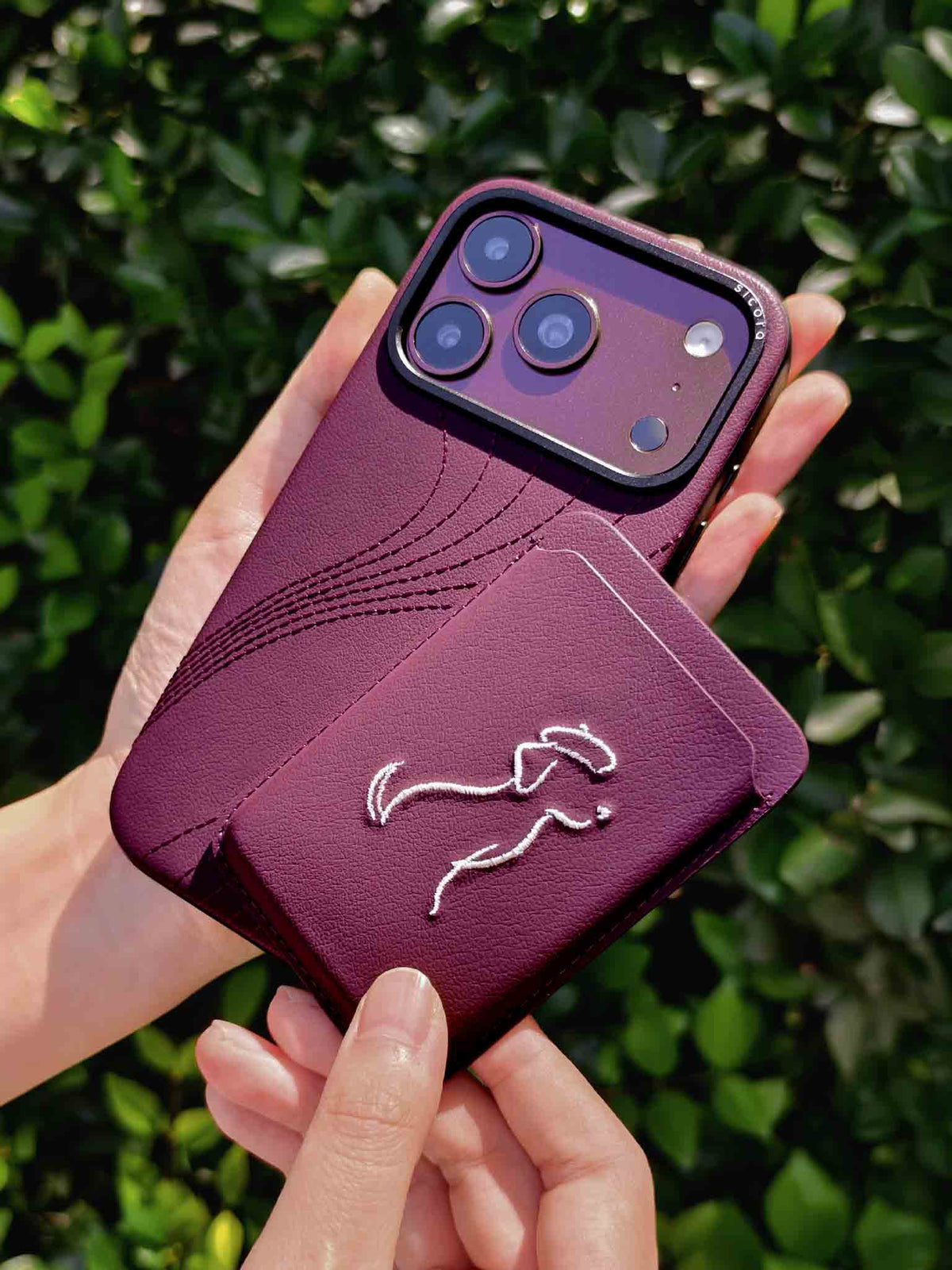 Hands holding a sicora vegan leather graphic magnetic card wallet in cherry purple with cream white embroidered dog line-drawing & a sicora iPhone 18 pro case