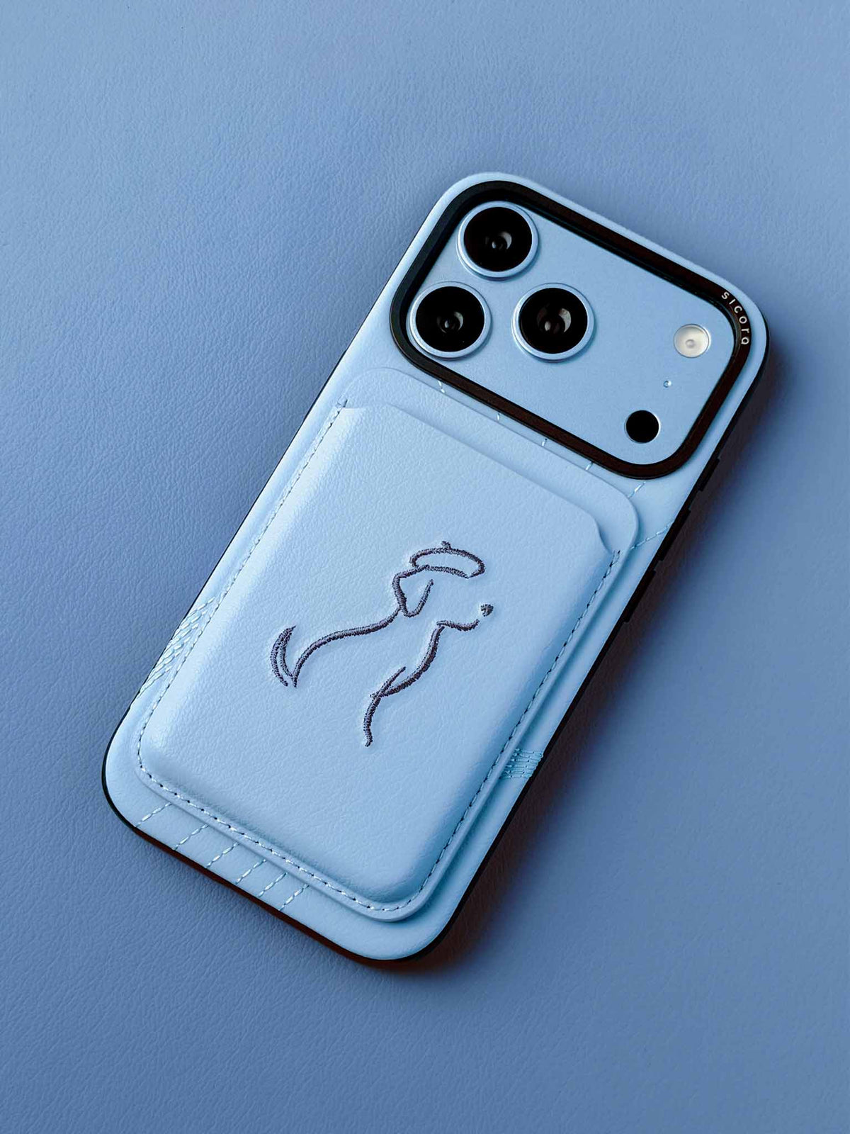 sicora vegan leather graphic magnetic card wallet in sky blue with grey blue embroidered dog line-drawing, on an iPhone 18 pro case