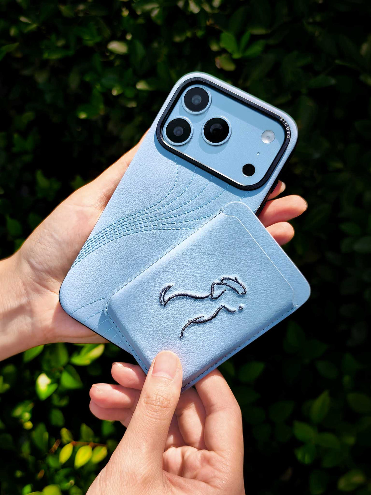 Hands holding a sicora vegan leather graphic magnetic card wallet in sky blue with grey blue embroidered dog line-drawing & a sicora iPhone 18 pro case
