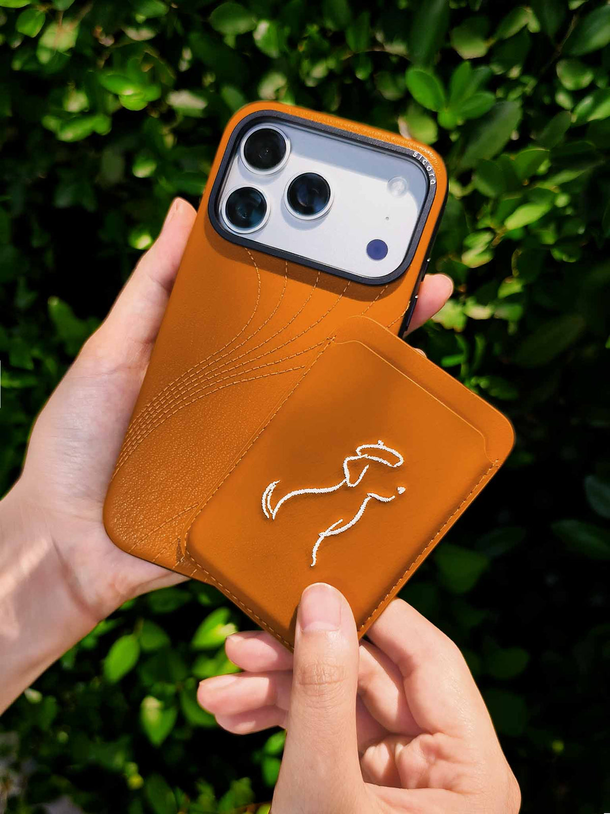 Hands holding a sicora vegan leather graphic magnetic card wallet in toffee gold brown with cream white embroidered dog line-drawing & a sicora iPhone 18 pro case