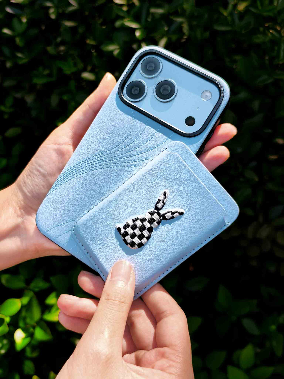 Hands holding a sicora vegan leather graphic magnetic card wallet in light blue with black and white embroidered rabbit artwork & a sicora iPhone 18 pro case