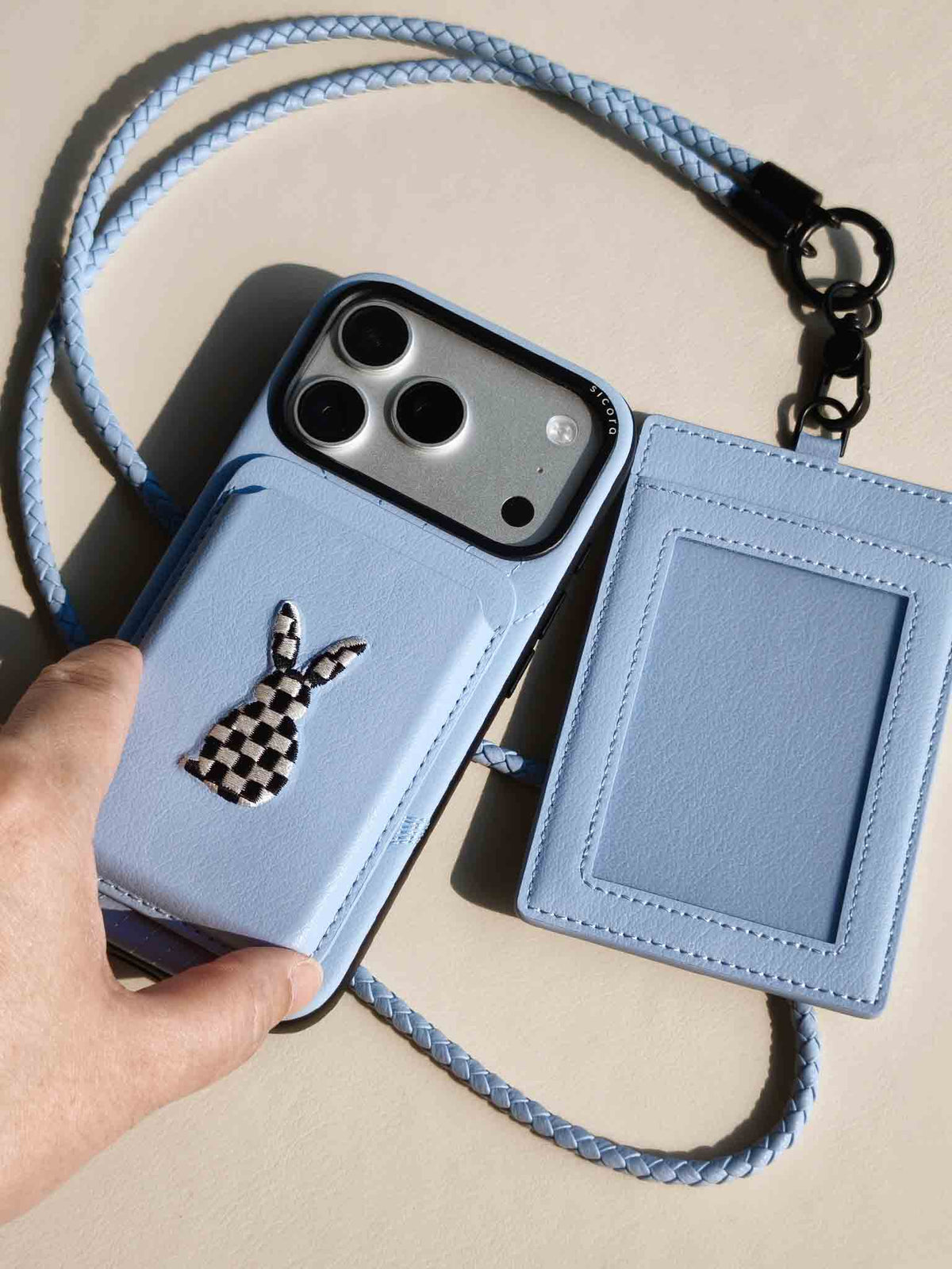 Hand holding a sicora vegan leather graphic magnetic card wallet in sky blue with black and white embroidered rabbit artwork, matching sicora iPhone case and ID badge holder with lanyard
