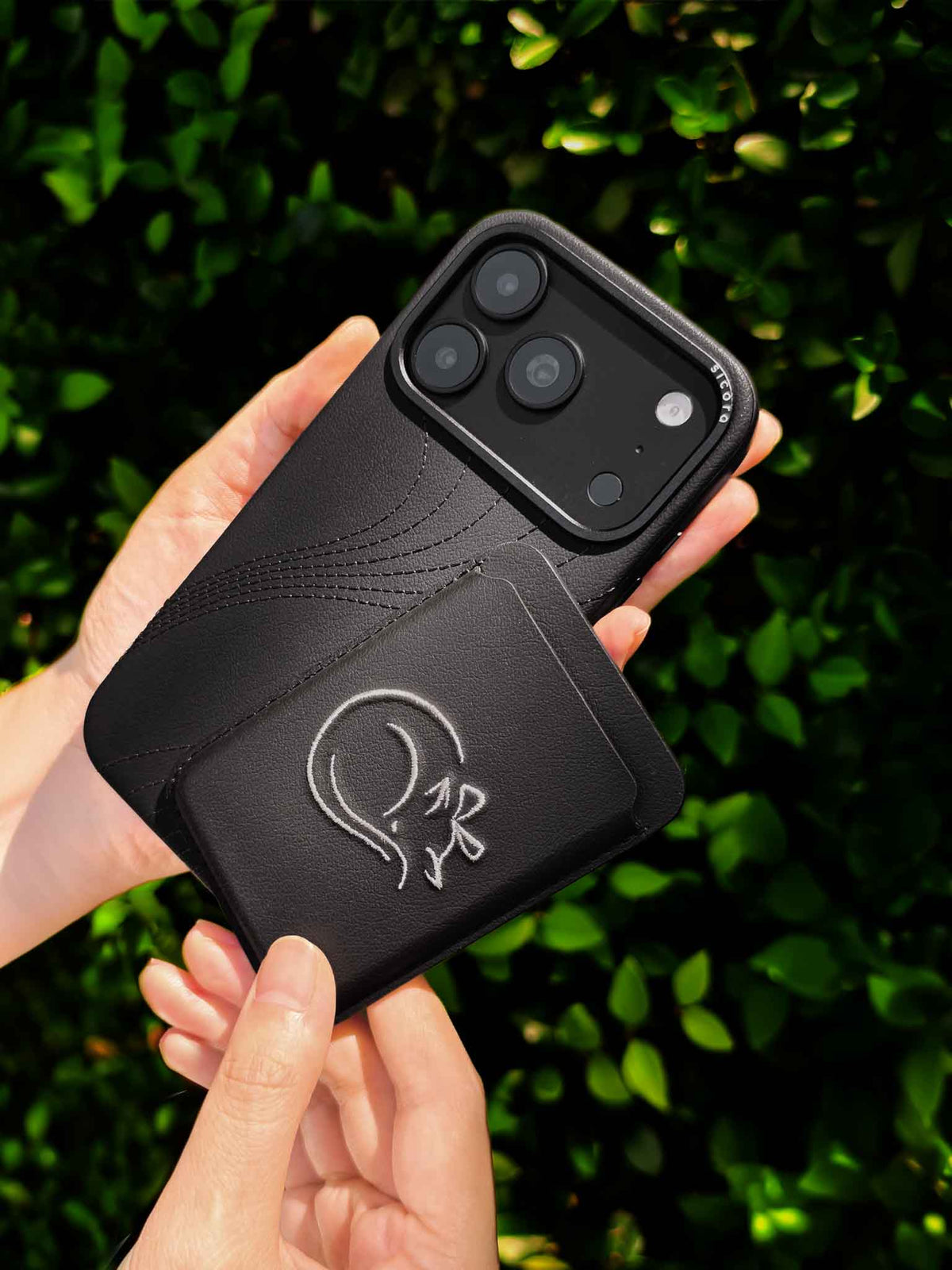 Hands holding a sicora vegan leather graphic magnetic card wallet in black with cream white embroidered cat line-drawing & a sicora iPhone 18 pro case