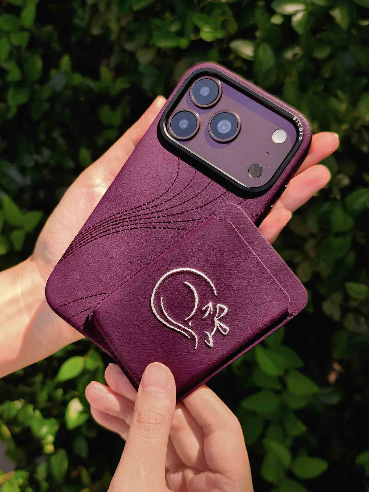Hands holding a sicora vegan leather graphic magnetic card wallet in cherry purple with cream white embroidered cat line-drawing & a sicora iPhone 18 pro case