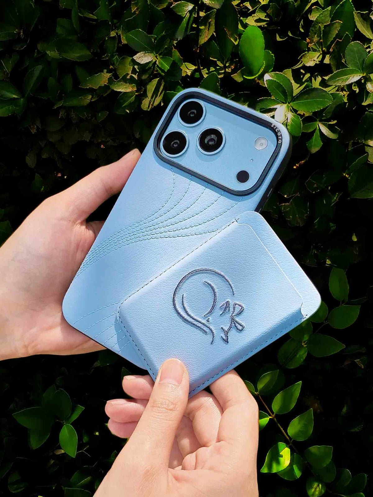 Hands holding a sicora vegan leather graphic magnetic card wallet in sky blue with grey blue embroidered cat line-drawing & a sicora iPhone 18 pro case