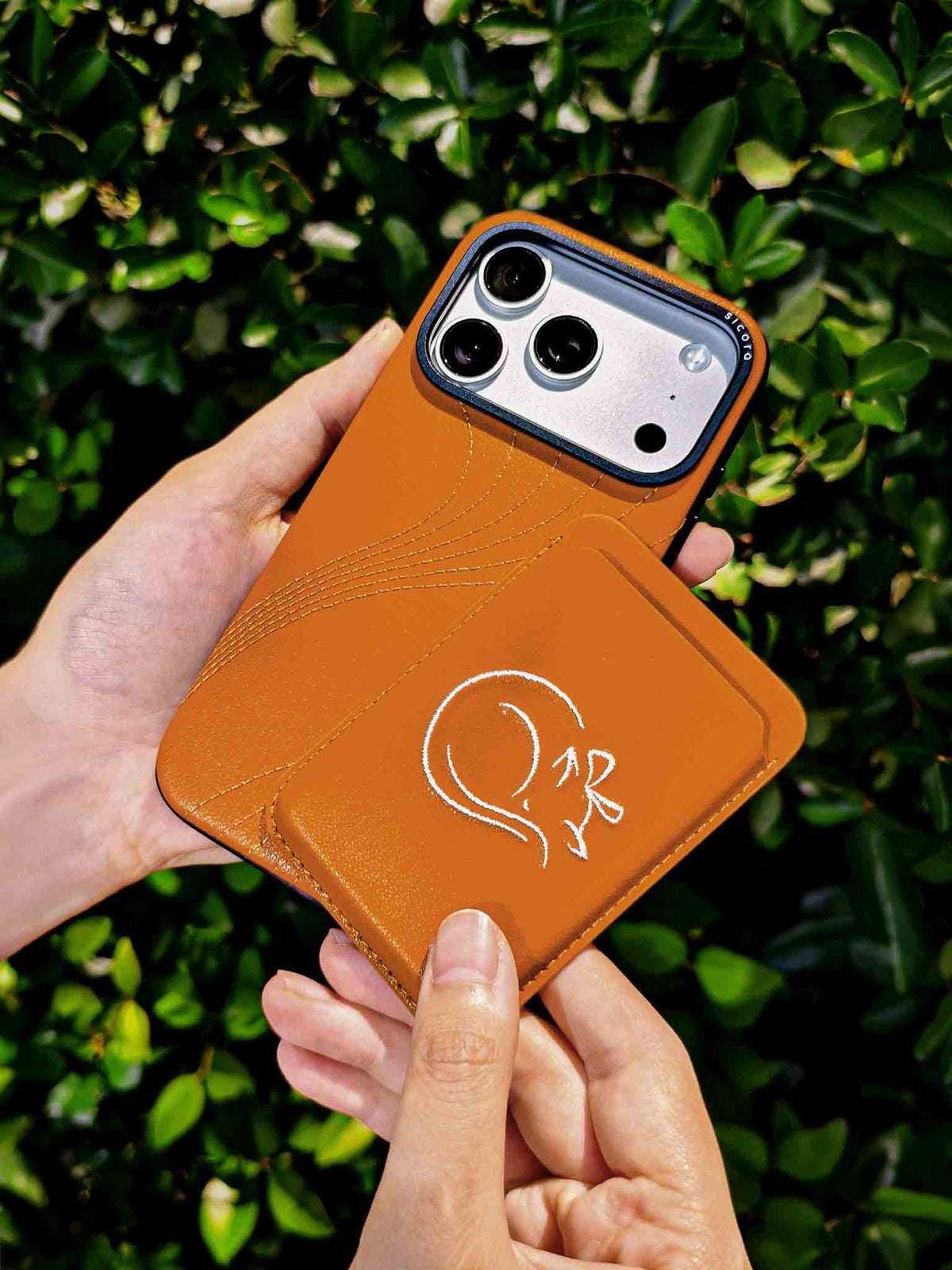 Hands holding a sicora vegan leather graphic magnetic card wallet in toffee gold brown with cream white embroidered cat line-drawing & a sicora iPhone 18 pro case