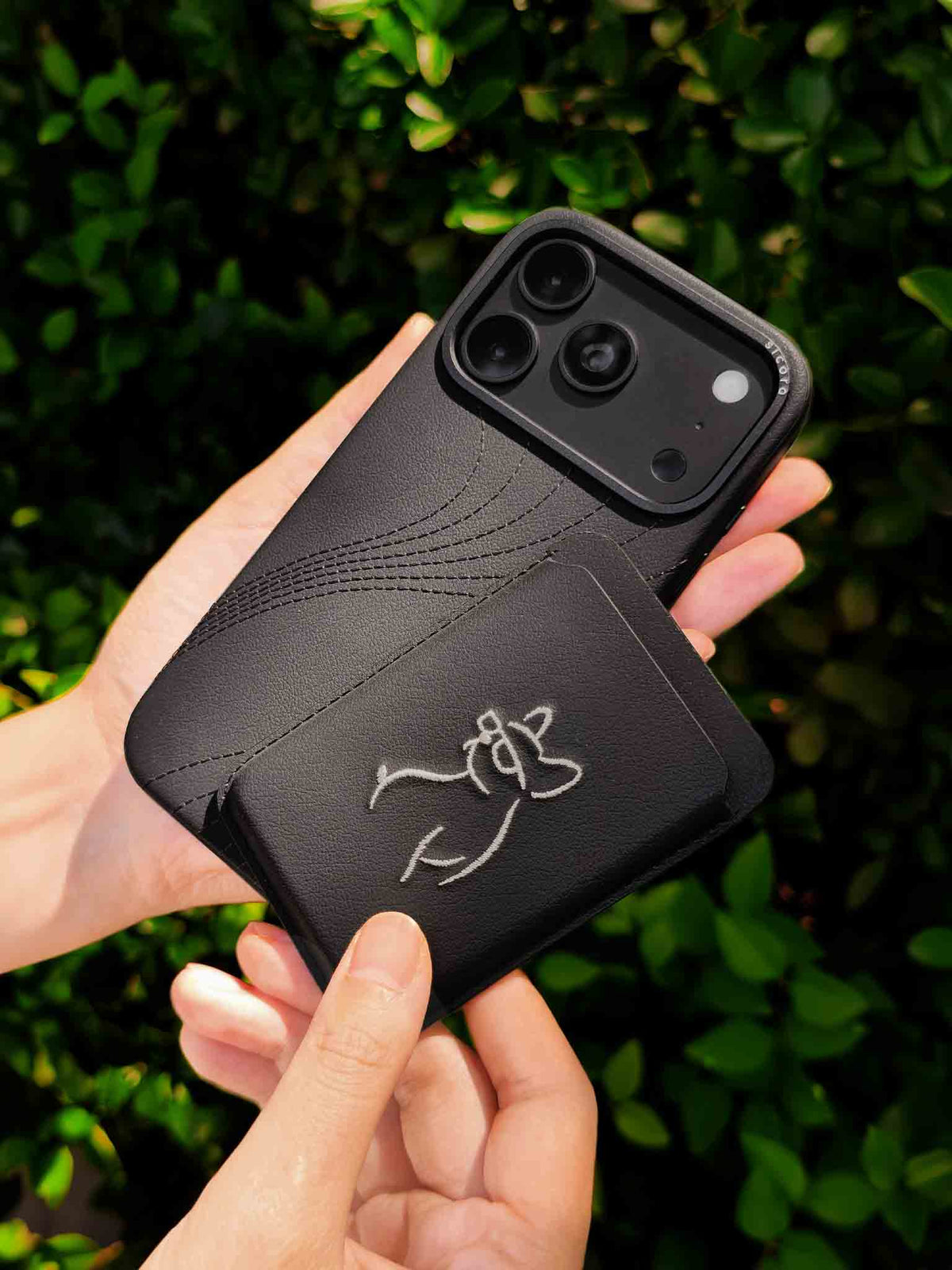 Hands holding a sicora vegan leather graphic magnetic card wallet in black with cream white embroidered dog line-drawing & a sicora iPhone 18 pro case