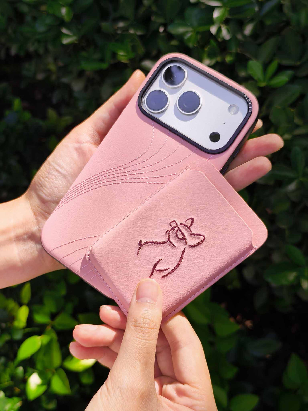 Hands holding a sicora vegan leather graphic magnetic card wallet in pink with dark burgundy embroidered dog line-drawing & a sicora iPhone 18 pro case