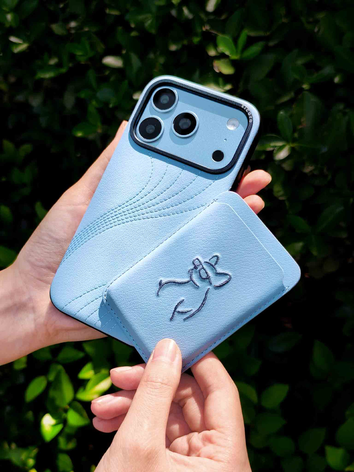 Hands holding a sicora vegan leather graphic magnetic card wallet in sky blue with grey blue embroidered dog line-drawing & a sicora iPhone 18 pro case