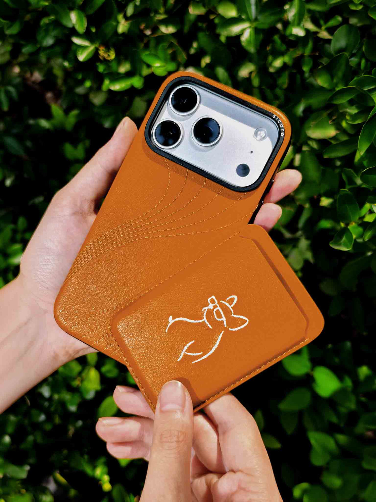 Hands holding a sicora vegan leather graphic magnetic card wallet in toffee gold brown with cream white embroidered dog line-drawing & a sicora iPhone 18 pro case