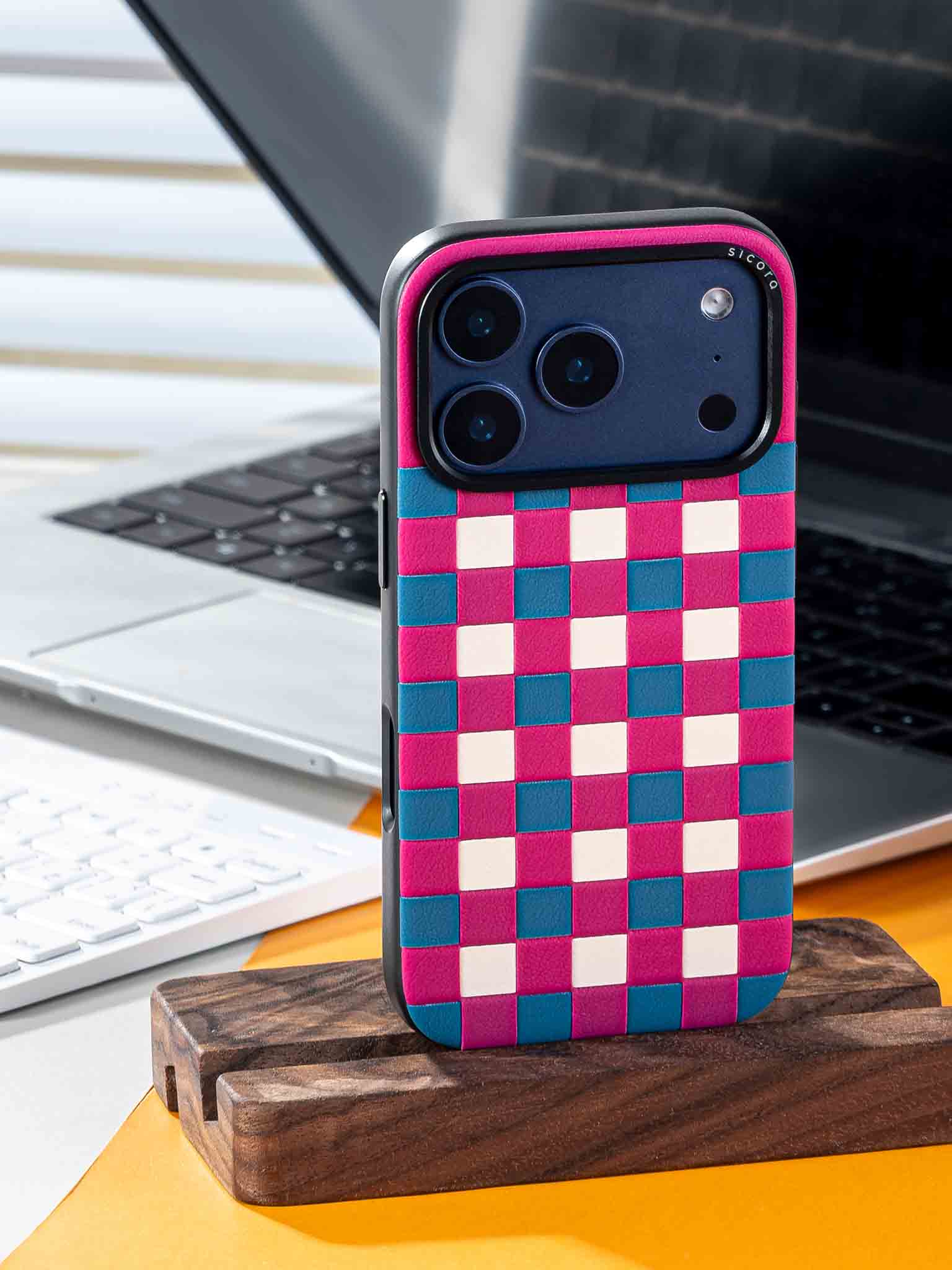 Sicora checkerboard phone case with magenta, sea blue and cream white checkered pattern on a wooden stand in front of a laptop