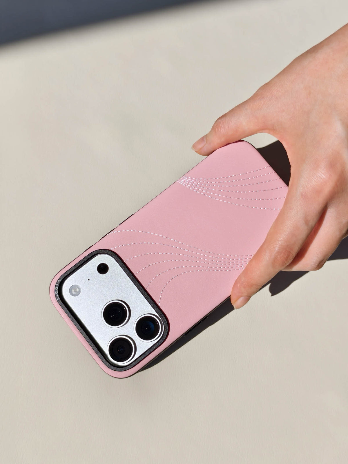 Hand holding a sicora pink iPhone 18 pro / iPhone 17 pro vegan leather phone case, flow case featuring tone-on-tone curvy embroidered stitch-lines on premium SILIO silicone vegan leather.