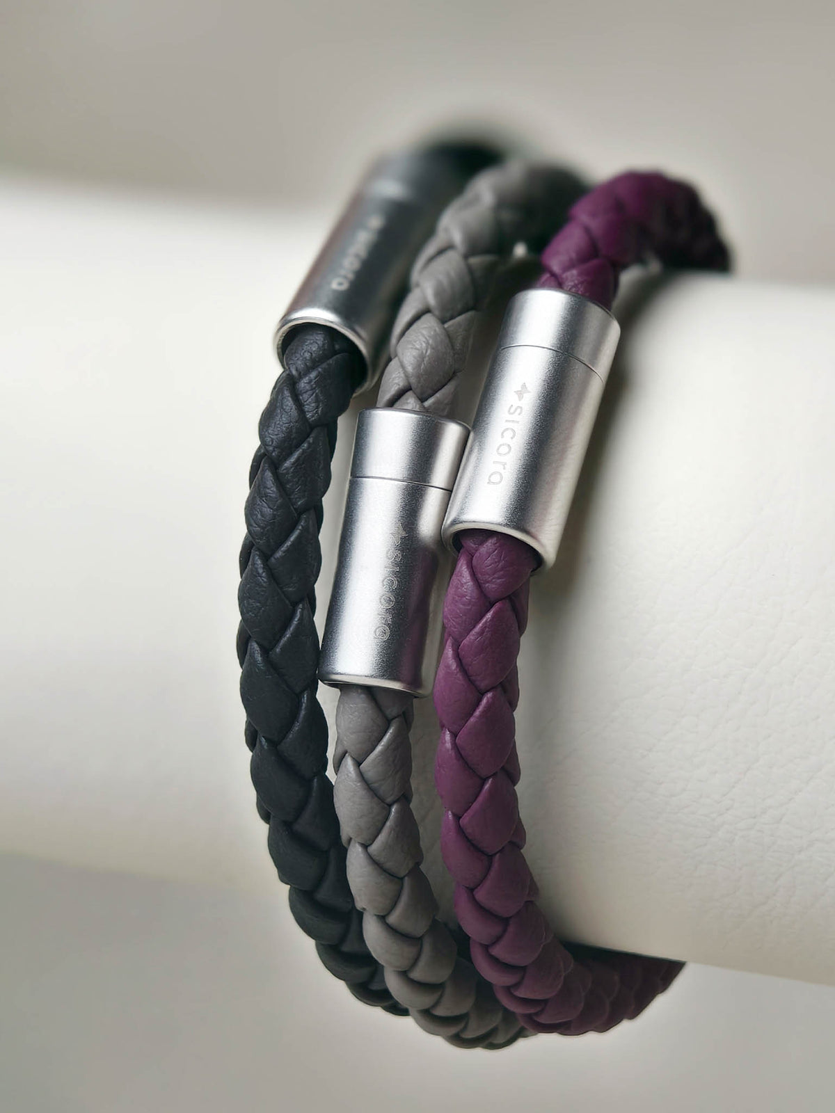 Three sicora braided leather bracelets in black, gray, and purple, each with a stainless steel cylindrical magnetic clasp, displayed on a white cylindrical stand.