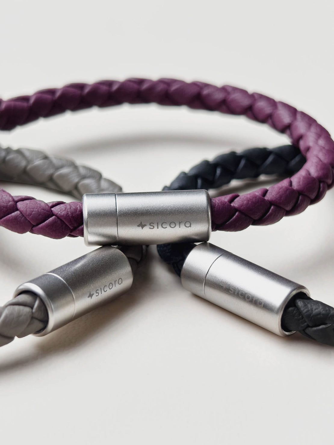 Three sicora braided leather bracelets with stainless steel magnetic clasps engraved with the 'sicora' brand name, arranged on a light gray background.