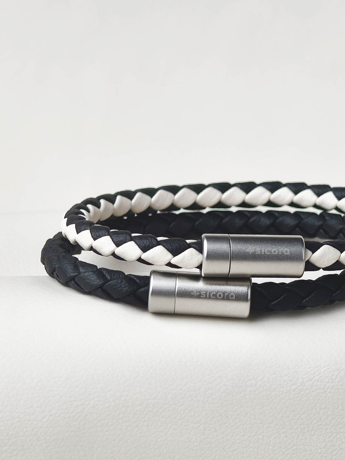 Sicora Black and white braided leather bracelets with stainless steel magnetic clasps engraved with the brand name 'sicora', displayed on a light gray textured surface.