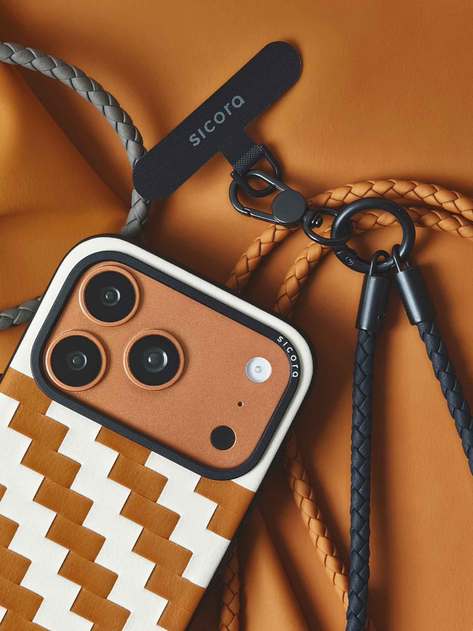 A close-up of an orange brown sicora zigzag iPhone 17 pro case with braided straps and black carabiner.