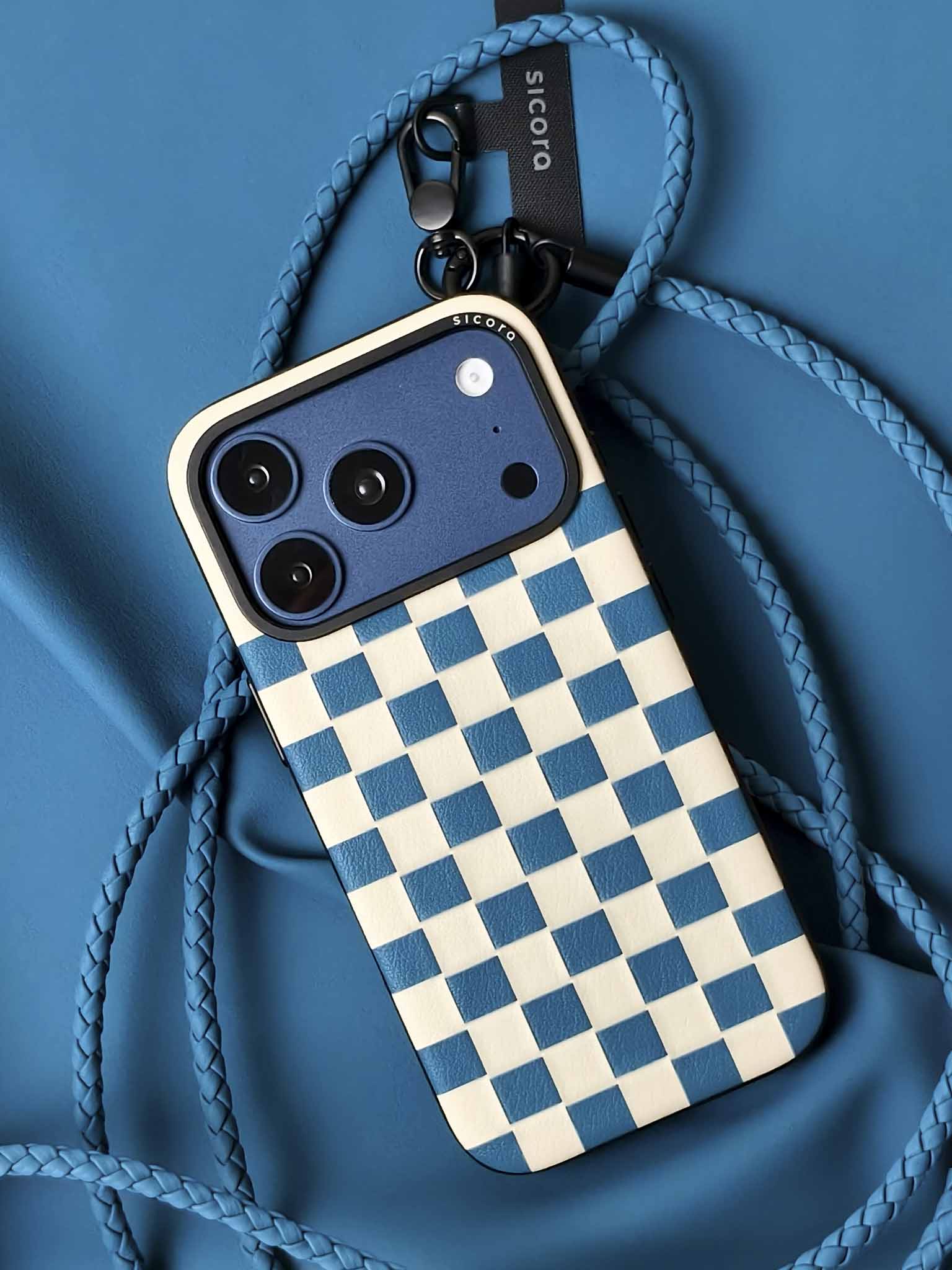 A blue sicora checkerboard iPhone 17 pro case with braided lanyards and black carabiner.