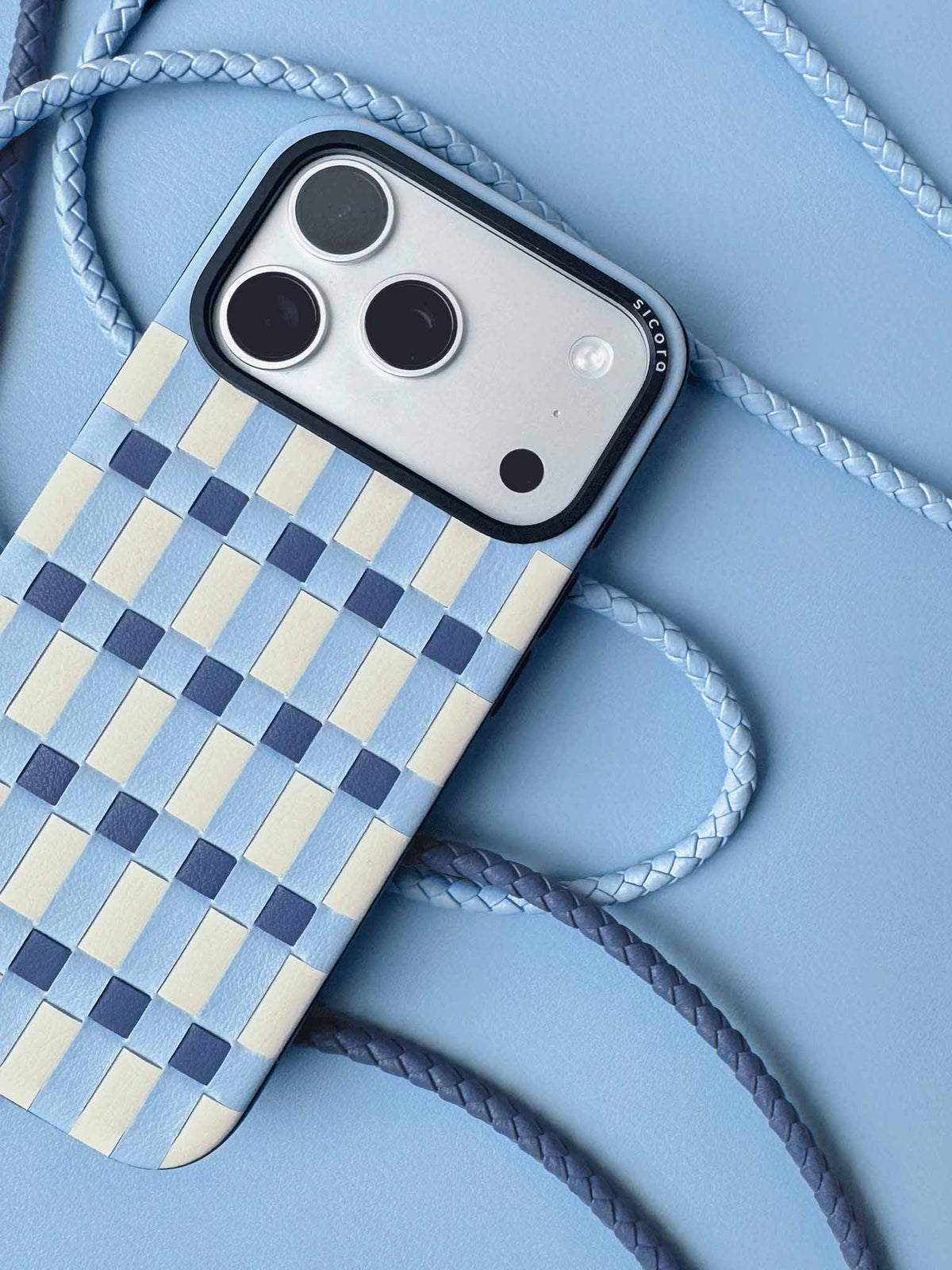 sicora handcrafted luxury iPhone 18 pro / iPhone 17 pro vegan leather phone case, pixel case featuring a woven / tiled pattern in sky blue, cream white and shadow blue geometric blocks, braided phone lanyards and blue leather background.